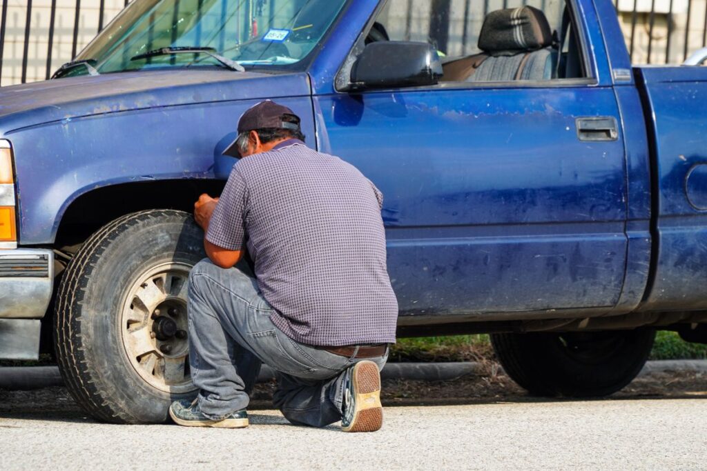 Driver kneeling to change a flat tire on a blue pickup truck at the roadside
