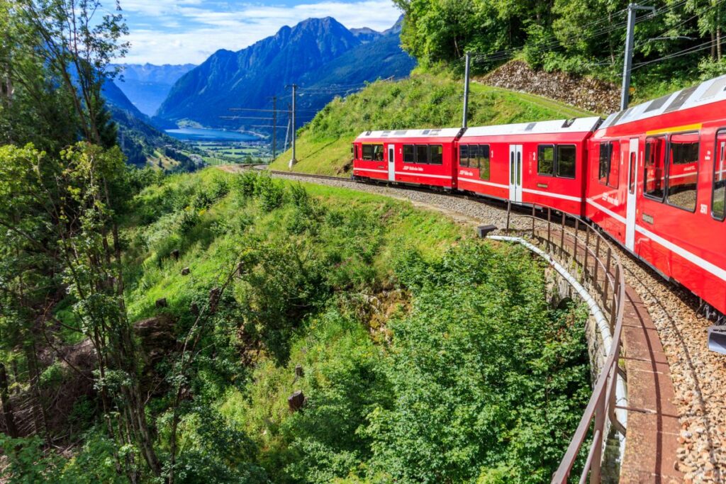 Red passenger train crossing a curved viaduct through green alpine mountains