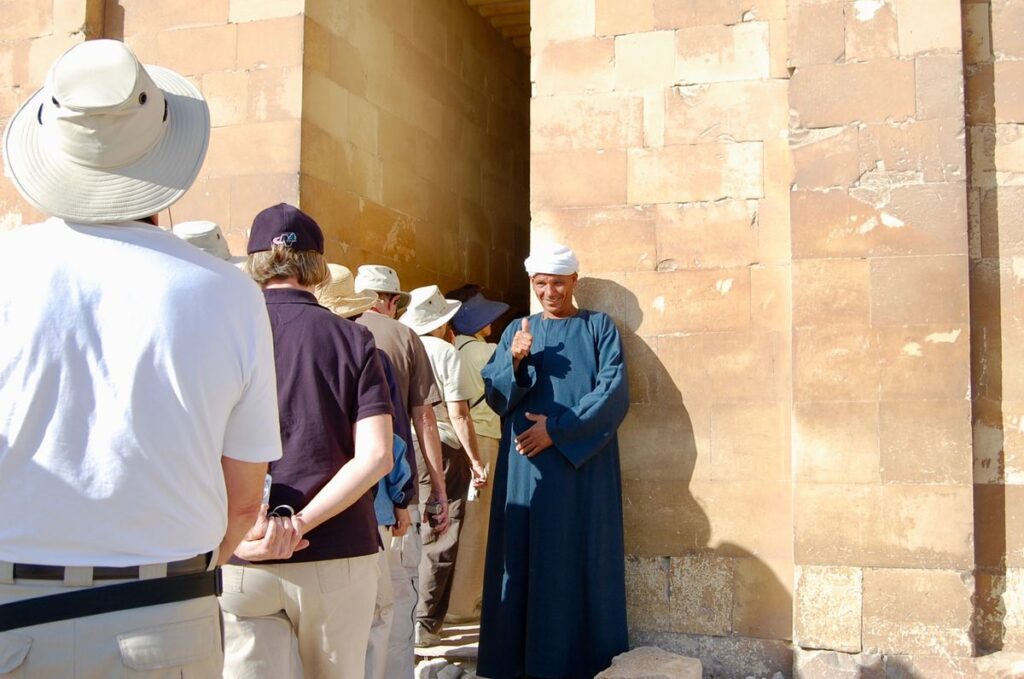 Group of older travelers in sun hats listening to a local guide beside an ancient stone wall