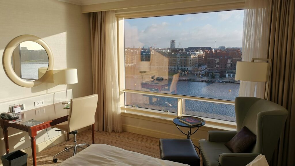 Hotellrum med skrivbord och fåtölj vid ett stort fönster med utsikt över vattnet
