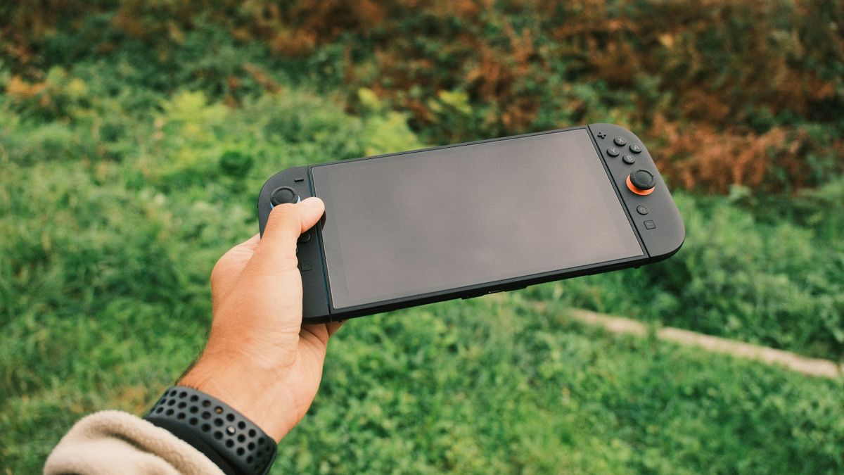 Hand hält eine Handheld-Konsole vor einer grünen Wiese